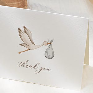 May include: A folded white card with a watercolor illustration of a stork carrying a bundle. The words "thank you" are written in a delicate script below the stork. The card is perfect for baby showers or new baby announcements.