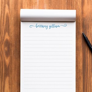 May include: A white notepad with blue lines and the name "brittany gilliam" written in blue cursive on the top. A black pen is lying on a wooden surface next to the notepad.