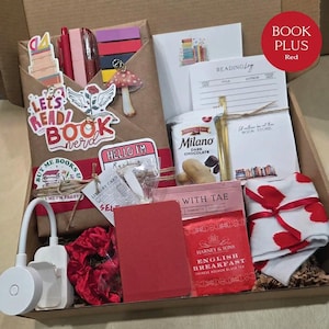 An open cardboard box filled with red-themed bookish merchandise, containing a wrapped book, red pens, sticky notes, floral stickers, a reading light, English breakfast tea, snacks, a red notebook, and red-patterned socks.