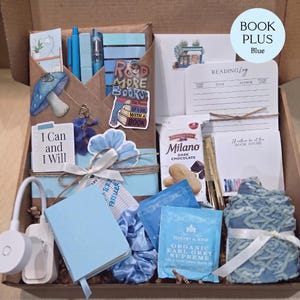 An open cardboard box filled with blue-themed bookish merchandise, including a book wrapped in brown paper, pens, sticky notes, a reading light, tea, snacks, a notebook, and socks.