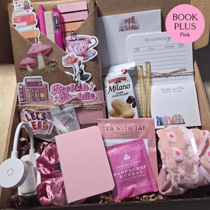 An open cardboard box filled with pink-themed bookish merchandise, including a wrapped book, pens, sticky notes, stickers, a reading light, tea, snacks, a notebook, and patterned socks.