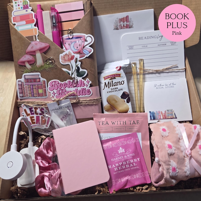 An open cardboard box filled with pink-themed bookish merchandise, including a wrapped book, pens, sticky notes, stickers, a reading light, tea, snacks, a notebook, and patterned socks.