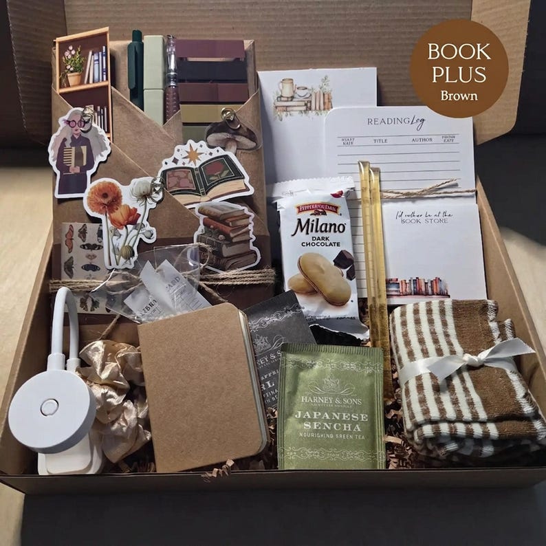 An open cardboard box filled with brown-themed bookish merchandise, featuring a wrapped book, pens, sticky notes, vintage floral and mushroom stickers, a reading light, green tea, cookies, a tan notebook, and brown striped socks.