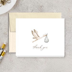 May include: A white card with a watercolor illustration of a stork carrying a bundle. The words "thank you" are written in a delicate script below the image. The card is paired with a cream-colored envelope.