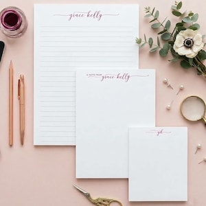 May include: Three white stationery pads of varying sizes with dark pink script text. The largest pad is lined and reads grace kelly. The medium pad reads A NOTE FROM grace kelly. The smallest pad reads gk. Includes rose gold pens and scissors.