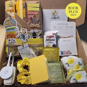 An open cardboard box filled with yellow-themed bookish merchandise, including a wrapped book, yellow pens, sticky notes, bee and floral stickers, a reading light, lemon tea, cookies, a yellow notebook, and lemon-patterned socks.