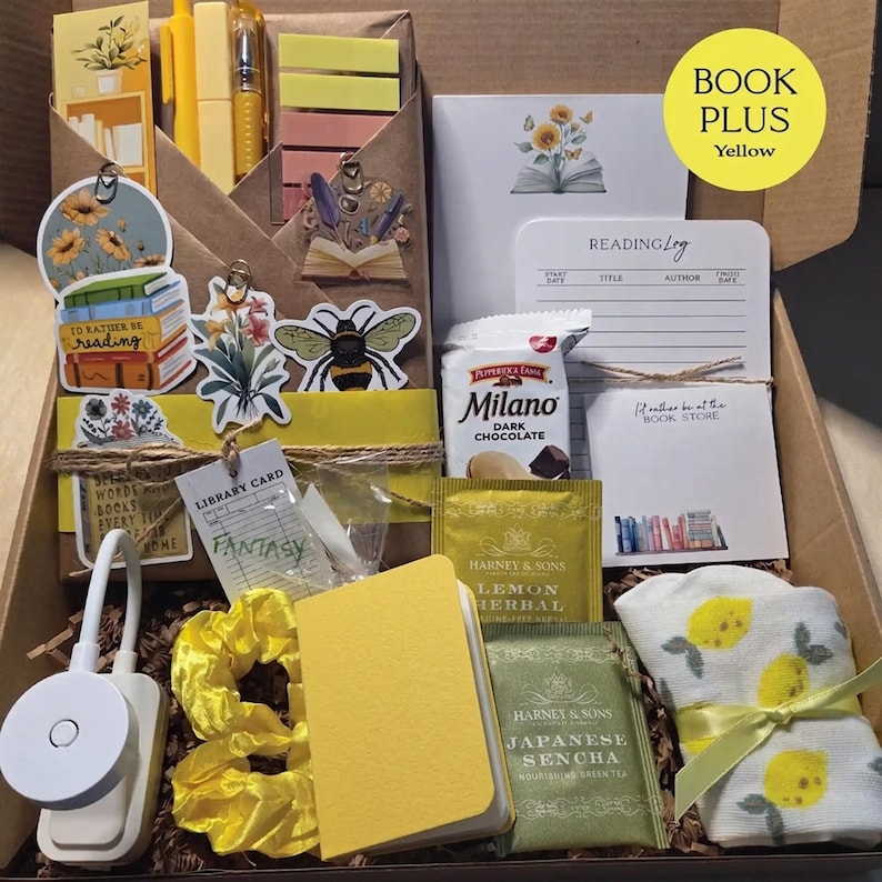 An open cardboard box filled with yellow-themed bookish merchandise, including a wrapped book, yellow pens, sticky notes, bee and floral stickers, a reading light, lemon tea, cookies, a yellow notebook, and lemon-patterned socks.