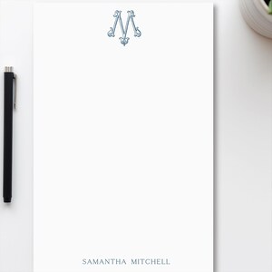 May include: White stationery with a blue decorative initial "M" at the top and the name "SAMANTHA MITCHELL" printed at the bottom. A black pen is on the left side of the stationery.