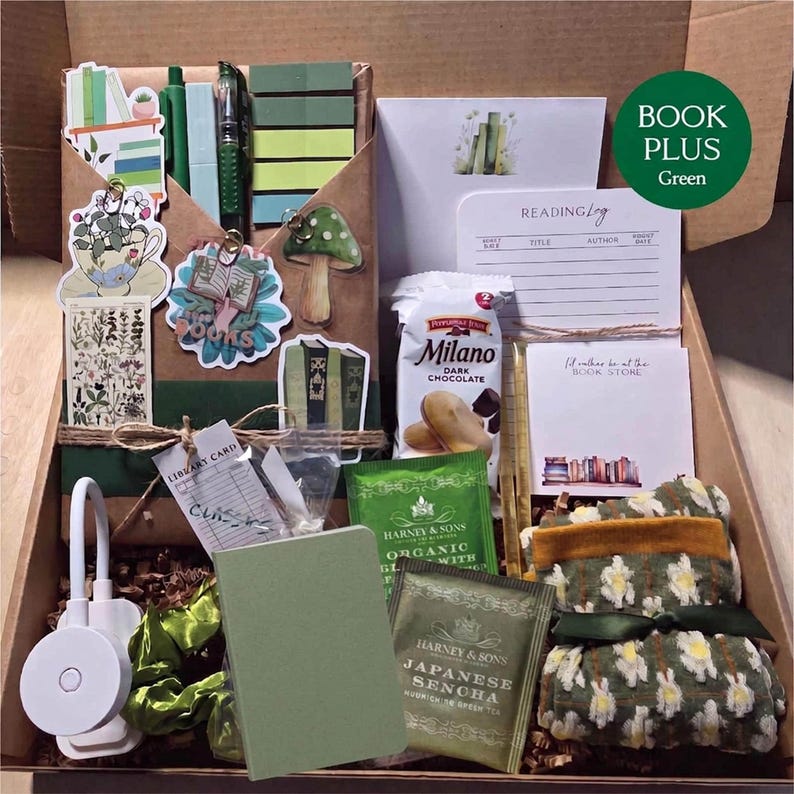 An open cardboard box filled with green-themed bookish merchandise, displaying a wrapped book, green pens, sticky notes, botanical stickers, a reading light, green tea, snacks, a green notebook, and patterned socks.