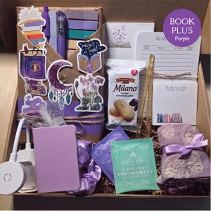 An open cardboard box filled with purple-themed bookish merchandise, featuring a wrapped book, purple pens, sticky notes, decorative stickers, a reading light, peppermint tea, snacks, a purple notebook, and a scarf.