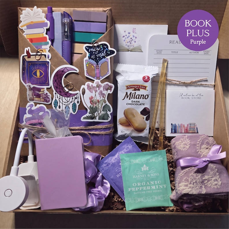 An open cardboard box filled with purple-themed bookish merchandise, featuring a wrapped book, purple pens, sticky notes, decorative stickers, a reading light, peppermint tea, snacks, a purple notebook, and a scarf.