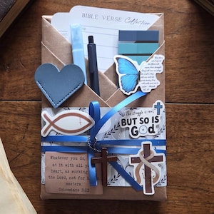 May include: A brown paper envelope containing a Bible Verse Collection card, a light blue highlighter, a black pen, blue sticky tabs, a blue heart cutout, and stickers featuring a blue butterfly, a fish, a cross, and text.