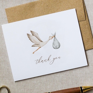 May include: A white card with a watercolor illustration of a stork carrying a bundle. The stork is light brown and gray, with a pink beak. The words "thank you" are written in a delicate script below the illustration. A brown envelope is behind the card.