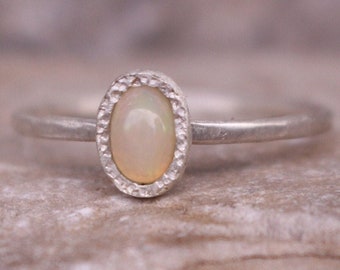 May include: A silver ring featuring an oval opal gemstone. The opal is a creamy white hue, set within a textured bezel. The ring's band is smooth, highlighting the gemstone's beauty.