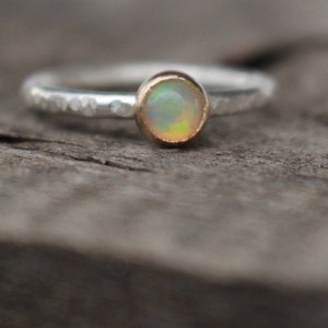 Peut inclure: Une bague en argent avec un anneau martelé et une petite pierre d'opale ronde sertie dans une lunette en or. L'opale affiche un mélange de couleurs, dont le blanc, le jaune et le vert. La bague est posée sur une surface en bois vieilli.