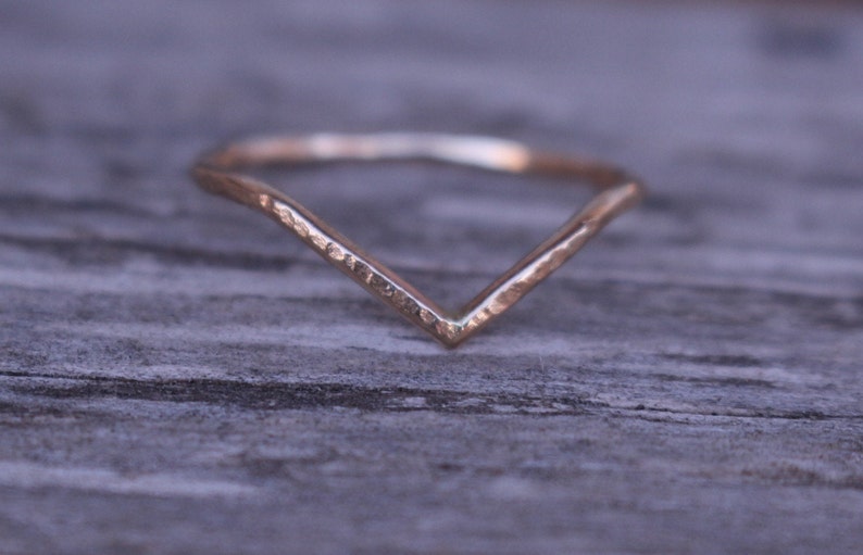 May include: A gold-coloured V-shaped ring. The ring has a hammered texture and a simple, elegant design. The ring is set against a blurred wooden background, highlighting the jewellery.