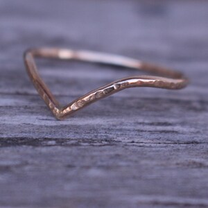 May include: A delicate, gold-coloured ring with a V-shaped design. The ring has a hammered texture and a subtle shine, suggesting a handcrafted piece. The ring is likely made of gold or a gold-toned metal.