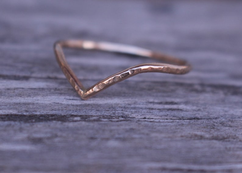 May include: A delicate, gold-coloured ring with a V-shaped design. The ring has a hammered texture and a subtle shine, suggesting a handcrafted piece. The ring is likely made of gold or a gold-toned metal.