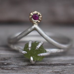 Pode incluir: Um anel de prata com um design único. O anel apresenta uma pedra verde clara, de corte bruto, presa por garras de prata. Um pequeno detalhe dourado com uma pedra preciosa vermelha fica no topo do design em forma de V do anel.