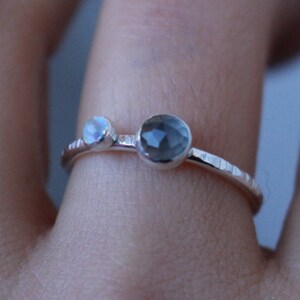 May include: A delicate silver ring featuring two gemstones. One is a small, iridescent moonstone, and the other is a larger, faceted, smoky gray stone. The band has a textured, hammered appearance. This ring is a piece of fine jewelry.