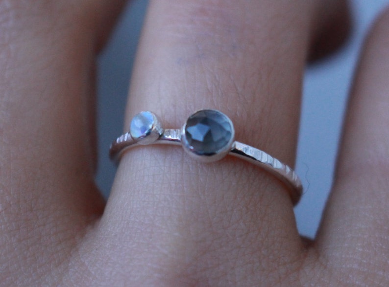 May include: A delicate silver ring featuring two gemstones. One is a small, iridescent moonstone, and the other is a larger, faceted, smoky gray stone. The band has a textured, hammered appearance. This ring is a piece of fine jewelry.