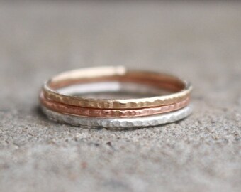 May include: A stack of three hammered metal rings in gold, rose gold, and silver. The rings are stacked together, with the gold ring on top, the rose gold in the middle, and the silver ring at the bottom. The rings have a textured surface.