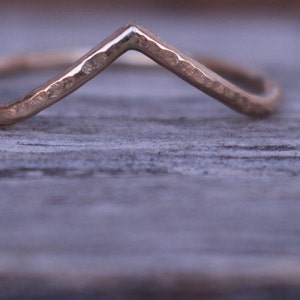 May include: A gold-coloured ring with a V-shaped design. The ring has a hammered texture and sits on a weathered wooden surface. The ring's design is simple and elegant, suitable for everyday wear.