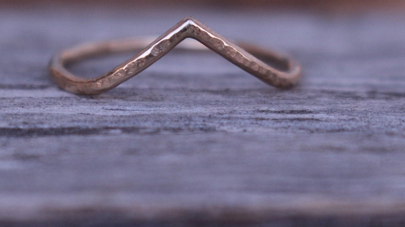 May include: A gold-coloured ring with a V-shaped design. The ring has a hammered texture and sits on a weathered wooden surface. The ring's design is simple and elegant, suitable for everyday wear.