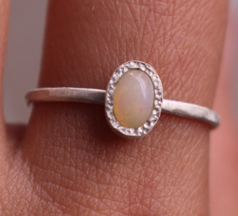 Puede incluir: Anillo de plata con una piedra de &oacute;palo ovalada. El &oacute;palo es de color blanco cremoso con una sutil iridiscencia, engastado en un bisel texturizado. La fina banda pulida le da al anillo un aspecto delicado y elegante.