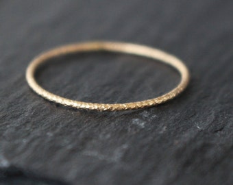 May include: A delicate gold ring with a textured surface. The ring is thin and circular, with a subtle, hammered appearance. The ring is set against a dark, textured background.