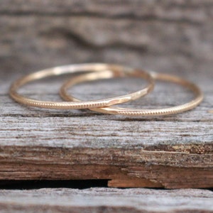Può includere: Due delicati anelli d'oro, uno con un motivo a pois strutturato e l'altro con un design sfaccettato. Gli anelli sono esposti su una superficie di legno invecchiato, mostrando i loro dettagli raffinati e il colore caldo.