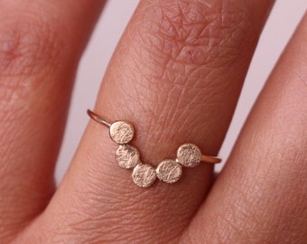 May include: A gold ring featuring a series of textured, circular elements arranged in an arc. The ring is worn on a finger, showcasing the delicate design and craftsmanship. The ring's warm gold color contrasts with the skin tone.