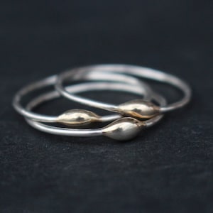 Op de afbeelding: Drie zilveren ringen met goudkleurige, druppelvormige accenten. De ringen zijn gestapeld en overlappen elkaar licht, tegen een donkere achtergrond. De sieraden zijn eenvoudig en elegant.