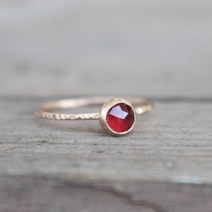 Puede incluir: Un anillo fino de oro con una pequeña piedra preciosa redonda y facetada de color rojo intenso engastada en un bisel. La banda del anillo tiene un acabado martillado y texturizado que capta la luz, contrastando con el engaste liso y pulido.