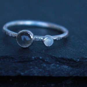 May include: A silver ring with two clear, round gemstones. The larger stone is on the left, and a smaller one is to the right. The band has a textured design. The ring is set against a dark background.
