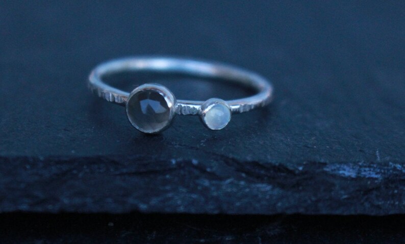 May include: A silver ring with two clear, round gemstones. The larger stone is on the left, and a smaller one is to the right. The band has a textured design. The ring is set against a dark background.