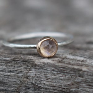May include: A delicate silver ring with a small, round gemstone set in a gold bezel. The gemstone has a pale, iridescent color. The ring is displayed on a weathered wooden surface, highlighting its simple elegance. Jewelry.