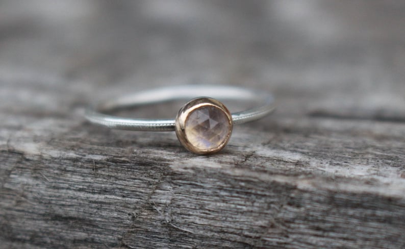 May include: A delicate silver ring with a small, round gemstone set in a gold bezel. The gemstone has a pale, iridescent color. The ring is displayed on a weathered wooden surface, highlighting its simple elegance. Jewelry.
