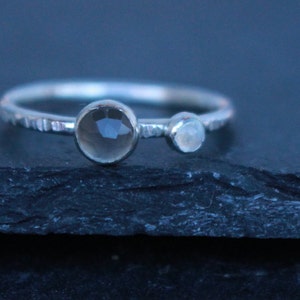 May include: A silver ring with two gemstones. The larger gemstone is a smoky quartz, and the smaller one is a moonstone. The band has a textured, hammered design. The ring is displayed on a dark, textured surface.