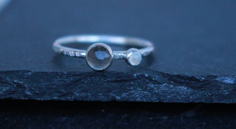 May include: A silver ring with two gemstones. The larger gemstone is a smoky quartz, and the smaller one is a moonstone. The band has a textured, hammered design. The ring is displayed on a dark, textured surface.