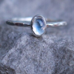Könnte beinhalten: Ein silberner Ring mit einem ovalen, schillernden Mondstein. Das Ringband hat eine gehämmerten Textur. Der Mondstein reflektiert blaues und weißes Licht, vor einem grauen Steinhintergrund. Ein Schmuckstück.