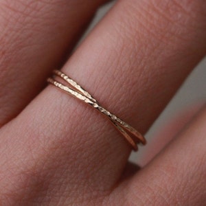 Könnte beinhalten: Nahaufnahme eines goldfarbenen Rings mit strukturierter Oberfläche. Der Ring hat ein Kreuz-Design, bei dem sich zwei Bänder kreuzen. Der Ring befindet sich an einem Finger und unterstreicht seinen zarten und eleganten Stil.