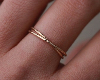 May include: Close-up of a gold ring with a textured finish. The ring has a crisscross design, with two bands crossing over each other. The ring is on a finger, highlighting its delicate and elegant appearance.