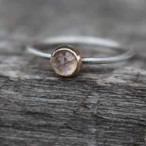 May include: A delicate silver ring with a small, round, faceted gemstone set in a gold bezel. The ring band has a textured design. The gemstone appears to be a light, translucent color, possibly a moonstone. The ring is displayed on a weathered wooden surface.