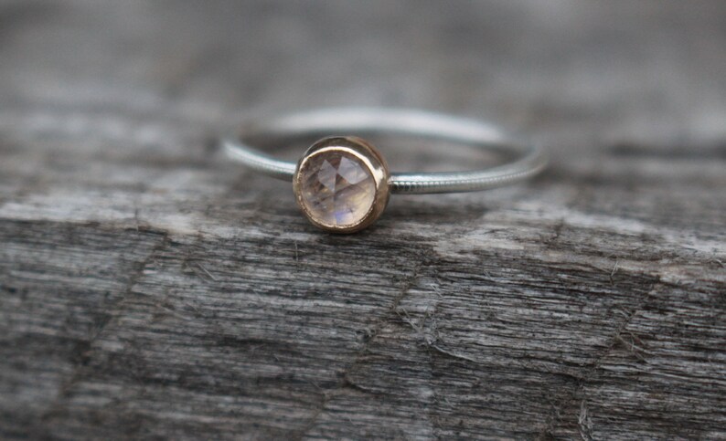 May include: A delicate silver ring with a small, round, faceted gemstone set in a gold bezel. The ring band has a textured design. The gemstone appears to be a light, translucent color, possibly a moonstone. The ring is displayed on a weathered wooden surface.