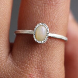 Puede incluir: Un anillo de plata con una piedra preciosa de &oacute;palo ovalada. El &oacute;palo est&aacute; engastado en un bisel texturizado, y la banda es fina y pulida. El anillo se muestra en un dedo, destacando su delicado dise&ntilde;o.