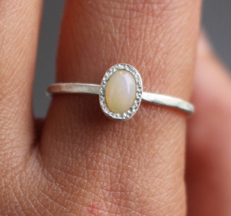 Puede incluir: Un anillo de plata con una piedra preciosa de &oacute;palo ovalada. El &oacute;palo est&aacute; engastado en un bisel texturizado, y la banda es fina y pulida. El anillo se muestra en un dedo, destacando su delicado dise&ntilde;o.