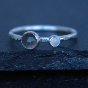 May include: A silver ring with two gemstones. The larger, faceted stone is a smoky gray color, and the smaller stone is a pale blue. The band has a textured, hammered appearance. Jewelry.