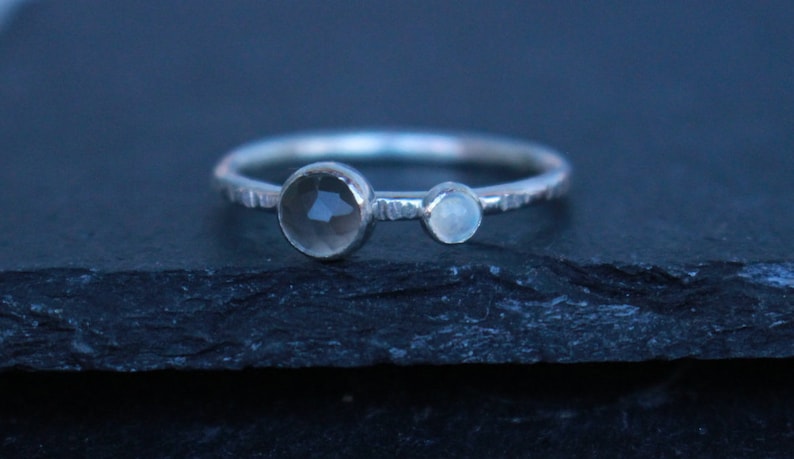 May include: A silver ring with two gemstones. The larger, faceted stone is a smoky gray color, and the smaller stone is a pale blue. The band has a textured, hammered appearance. Jewelry.