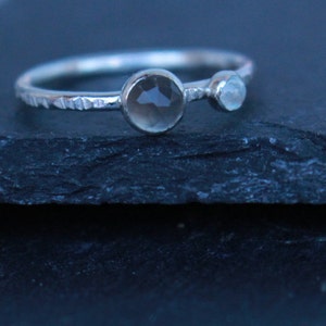 May include: A silver ring with a textured band features two gemstones: a larger, round, smoky quartz and a smaller, light blue stone. The ring is set against a dark, textured surface.